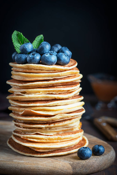 Closeup view of stack of homemade organic sweet pancakes or crepes decorated with fresh blueberries and mint leaves topping served on cutting board against black wall on kitchen table. Vertical - Фото, изображение