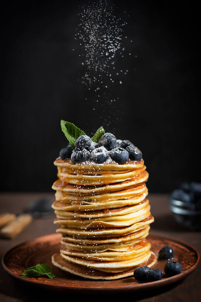 Pile of homemade organic sweet pancakes or crepes decorated with fresh blueberries and mint leaves topping served on plate against black wall on kitchen table with sugar powder sprinkles. Vertical - Фото, изображение
