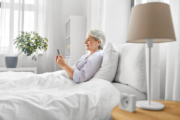 senior woman with smartphone and earphones in bed - Zdjęcie, obraz