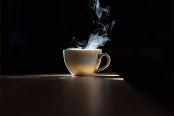 Horká voda v bílém šálku kávy na dřevěném stole s kouřem a přírodním světlem na černém pozadí - Fotografie, Obrázek