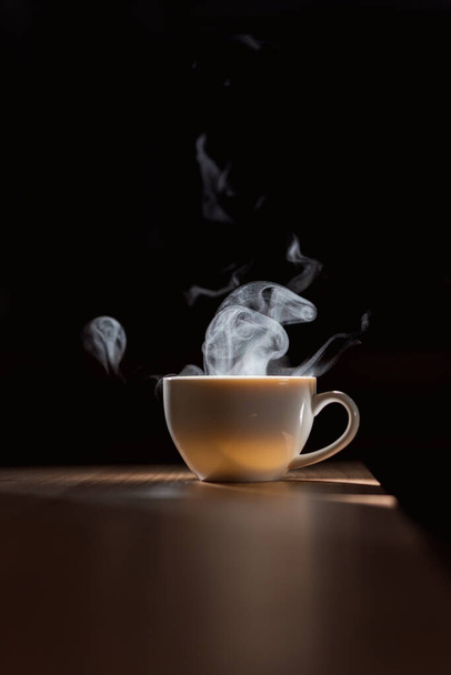 Agua caliente en taza de café blanco sobre mesa de madera con humo y luz natural sobre fondo negro - Foto, Imagen