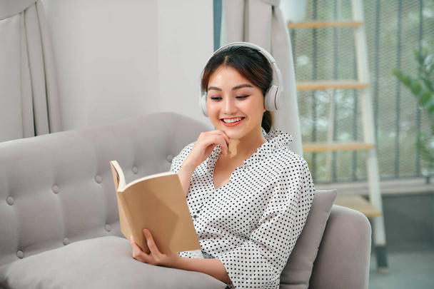 приваблива азіатська молода жінка відпочиває за допомогою смартфона для відтворення програми потокової передачі музики під час читання книги, що сидить на дивані або дивані в критому саду будинку
 - Фото, зображення