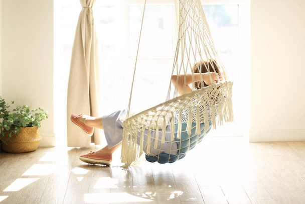 Femme rêveuse couchée dans une chaise suspendue et profitant d'un week-end dans une chambre confortable avec lumière du soleil - Photo, image