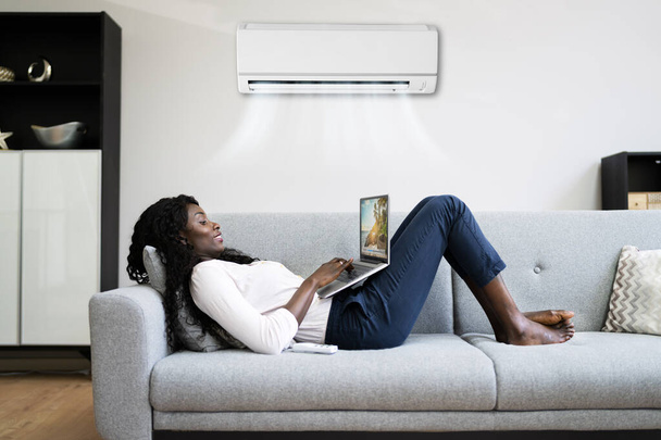 Young Happy Woman Lying Under Air Conditioner On Couch Using Laptop At Home - Foto, imagen