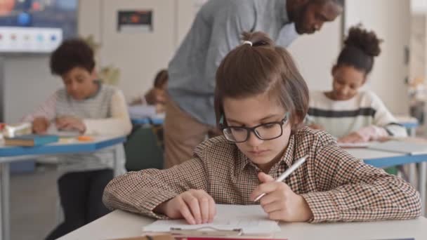 Waist-up slowmo van de Kaukasische 11-jarige schooljongen zitten aan het bureau in de klas doen van de test, terwijl jonge mannelijke Afro-Amerikaanse leraar hem te helpen door te wijzen op een stuk papier - Video