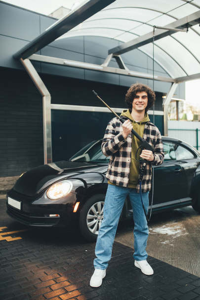 Усміхнений чоловік з мийкою під тиском стоїть біля авто на відкритому повітрі
  - Фото, зображення