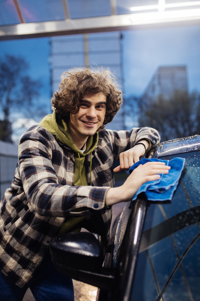 Göndör férfi mosolyog a kamerába, miközben nedves autót takarít a szabadban este  - Fotó, kép