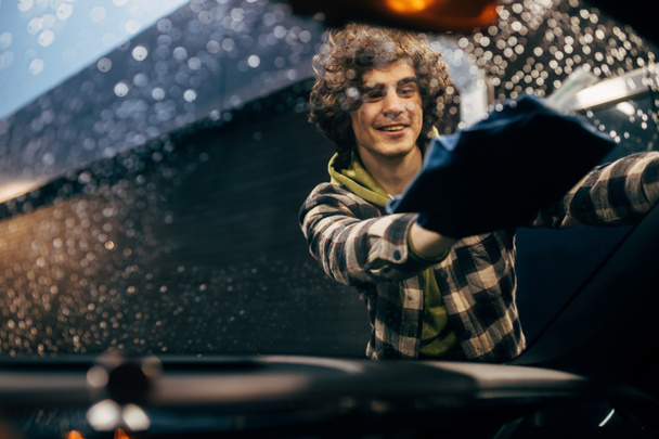 Conductor sonriente limpiando parabrisas de coche con trapo en la noche  - Foto, Imagen