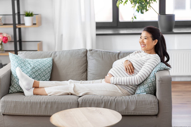 happy pregnant asian woman sitting on sofa at home - Foto, imagen
