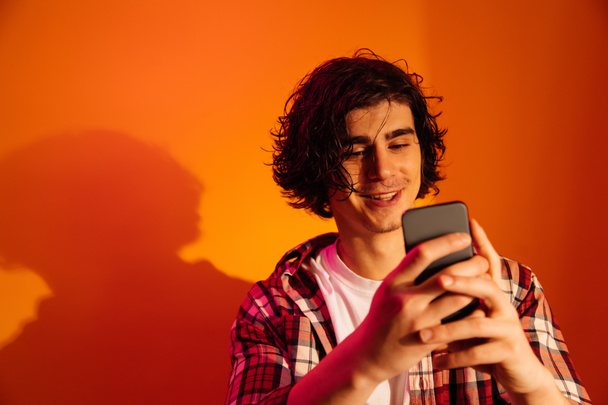 Young man smiling while using smartphone on orange background - Foto, Imagen