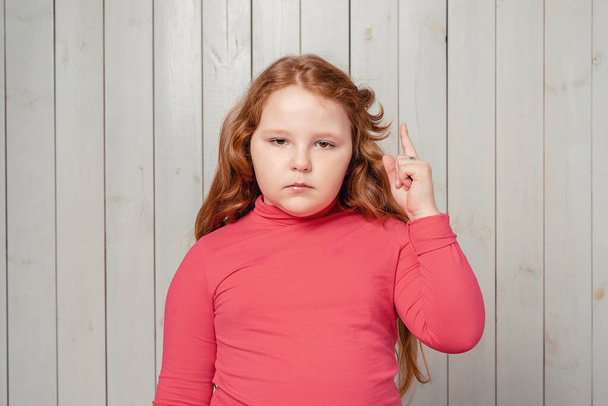 Portrait of cute redhead preteen girl, pointing with finger up giving advice. Studio shot, light wooden background - Foto, afbeelding