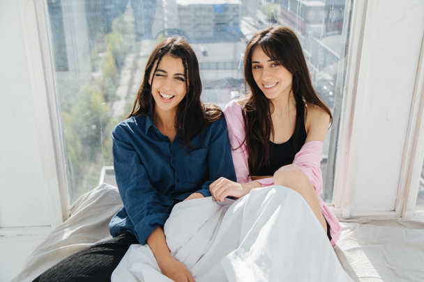 alegres mujeres armenias sonriendo a la cámara mientras se sienta cerca de la ventana en la ropa de cama blanca - Foto, Imagen