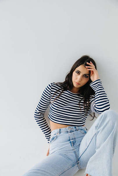 brunette armenian woman in striped pullover and jeans sitting on white - Fotografie, Obrázek