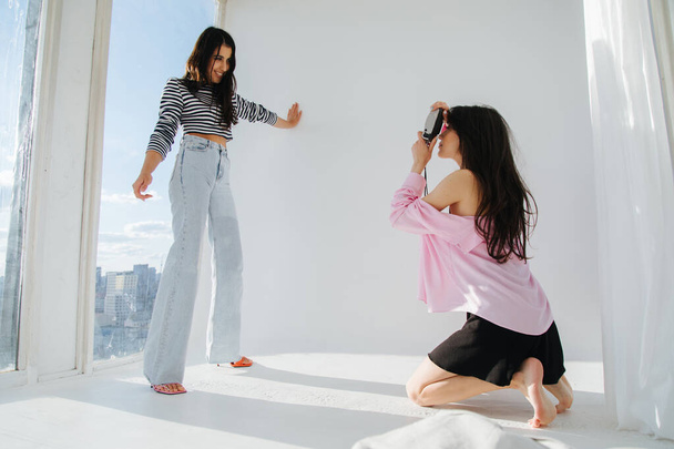 jovem mulher tirando foto de um amigo sorridente e na moda posando perto da janela em luz natural - Foto, Imagem