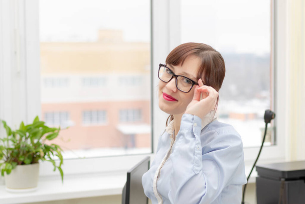 młoda ładna kobieta biznesu w okularach i niebieska bluzka stoi na biurku w biurze na tle okna światła - Zdjęcie, obraz