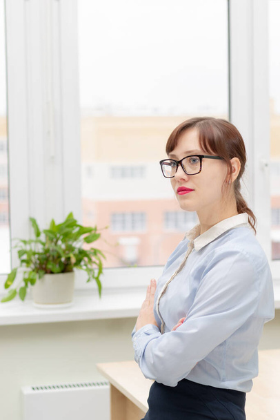 young pretty business woman in glasses and a blue blouse stands at the desktop in the office against the background of a light window - Фото, изображение