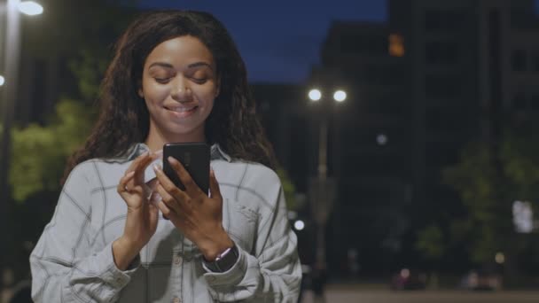 Urban Lifestylekonzept. Junge positive Afroamerikanerin textet auf Smartphone, steht im Nachtpark, freier Raum - Filmmaterial, Video