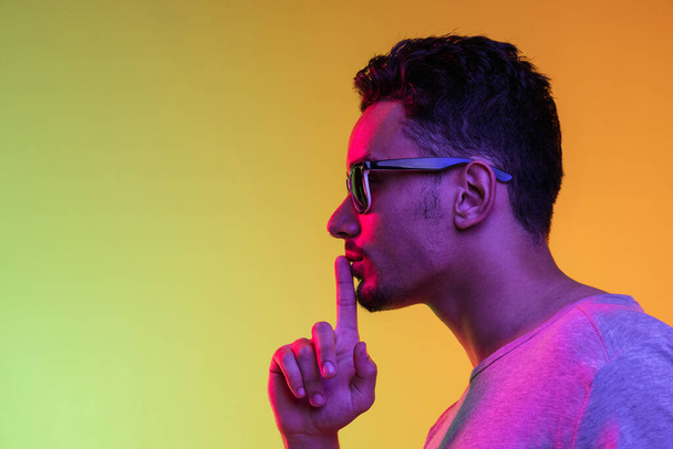 Portrait of Latina young man in sun glasses posing isolated on gradient yellow green background in neon light. Side view - Fotografie, Obrázek