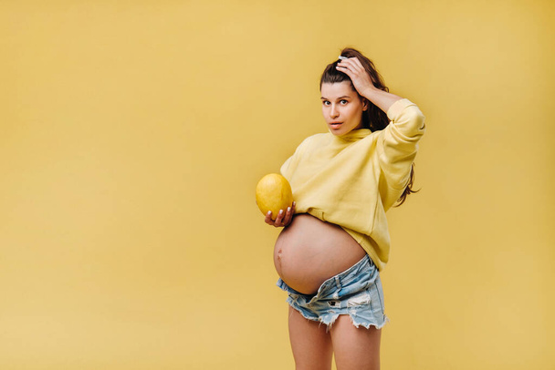 a pregnant girl with a yellow jacket stands with a melon in her hands on a yellow isolated background. - Photo, image