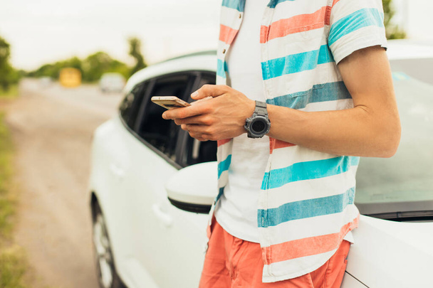 Handsome young man in summer clothes and sunglasses standing near his luxury white car and using smartphone, holding in hand, outdoors on a summer sunny day - Photo, Image