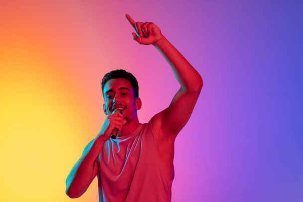 Portrait of Latina young man, singer with microphone performing isolated on gradient yellow purple background in neon light. Concept of emotions, facial expression - Foto, immagini