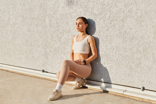 Athletic young woman sitting and leaning against gray wall looking away with calm and serious facial expression, wearing stylish sporty white top and beige leggins, healthy lifestyle, sport. - Photo, Image