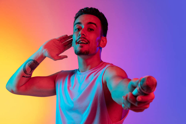 Portrait of Latina young man, singer with microphone performing isolated on gradient yellow purple background in neon light. Concept of emotions, facial expression - Fotografie, Obrázek