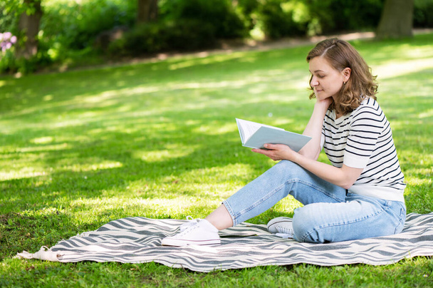 Beautiful young woman read book while relaxation in green big park at summertime - Photo, Image