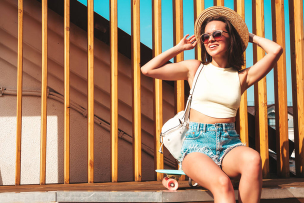 Portrait of young beautiful smiling hipster woman in trendy summer jeans shorts and hat. Sexy carefree model posing on the street background at sunset. Positive model outdoors with briefcase - Foto, Imagem