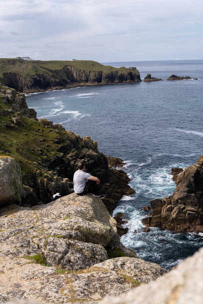 Foto eines jungen und attraktiven Mannes, der auf einem Felsen neben einer Klippe inmitten der Natur in Land 's End, Großbritannien, sitzt. Erstaunliche Landschaft an einem sonnigen Tag. - Foto, Bild