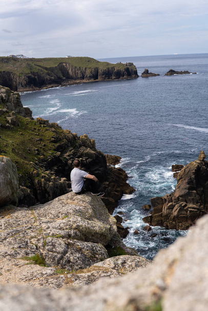 Φωτογραφία ενός νέου και ελκυστικού άνδρα που κάθεται σε ένα βράχο δίπλα σε ένα βράχο στη μέση της φύσης, στο Land 's End, Ηνωμένο Βασίλειο. Καταπληκτικό τοπίο σε μια ηλιόλουστη μέρα. - Φωτογραφία, εικόνα