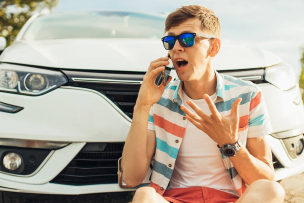 Excited screaming young man in sunglasses talking on mobile phone while sitting near his car on the road - Fotografie, Obrázek