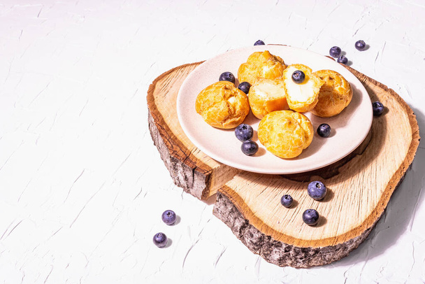 Profiteroles with blueberries. Custard cakes, Eclair, cream puff on a wooden stand, fashionable hard light, dark shadow. White plaster background, copy space - Photo, Image