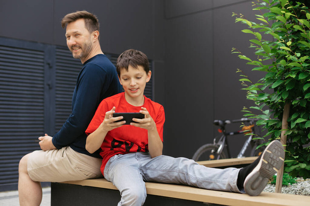 Papá e hijo entre las estructuras de hormigón utilizan dispositivos mod para pasar tiempo juntos, jugar videojuego en línea, concepto de crianza moderna - Foto, imagen