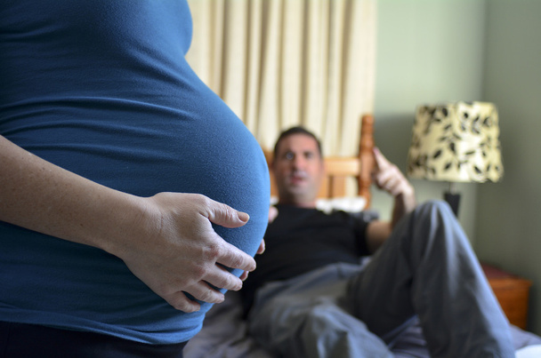 Pregnant woman holds her abdomen and blaming man lying on double bed in the background. Concept photo of pregnancy, pregnant woman lifestyle.copyspace - Foto, Bild