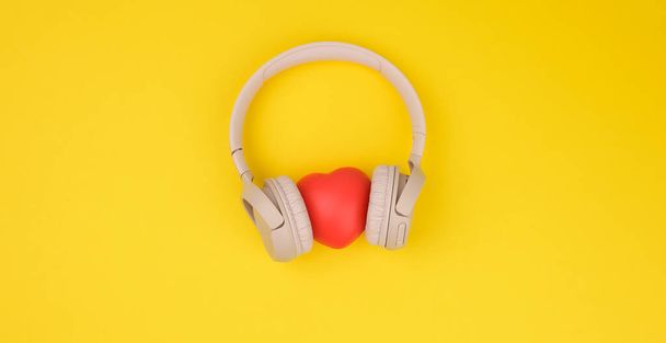 punainen sydän ja beige langattomat kuulokkeet keltaisella pohjalla. Top näkymä moderni gadget - Valokuva, kuva