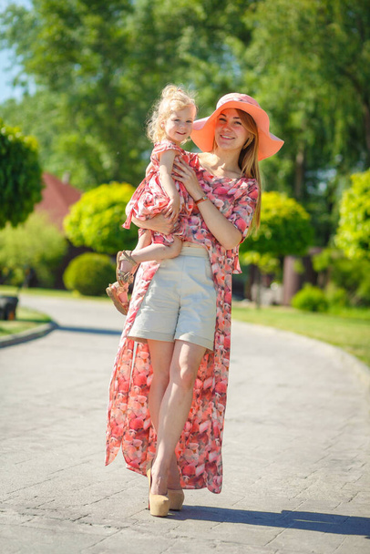 A charming girl in a light summer sundress walks in a green park with her little daughter, holding her in her arms. Enjoys warm sunny summer days. - Foto, Imagem