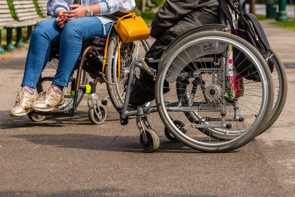 Pohled pro dvě ženy nohy sedí na invalidním vozíku. Dva lidé s invalidním vozíkem odpočívají v parku. Žena ve zlatých botách, džínách, žluté kabelce a bílém kabátě potkává další ženy v šedém a černém oblečení. - Fotografie, Obrázek