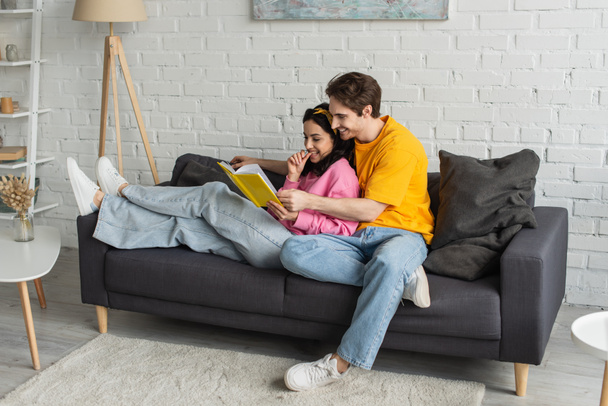 happy young couple resting on couch, hugging and holding book in living room - 写真・画像