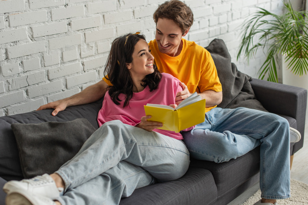 sourire jeune couple reposant sur le canapé, étreignant et tenant le livre dans le salon - Photo, image