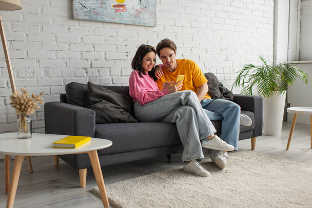 pareja joven y positiva sentada en el sofá y sosteniendo el teléfono celular en la sala de estar - Foto, Imagen