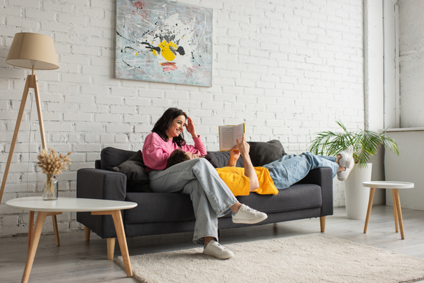 sorridente giovane donna seduta sul divano con fidanzato sdraiato e lettura libro in soggiorno - Foto, immagini