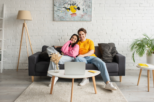 smiling young couple sitting on couch and watching movie on laptop in living room - Photo, Image