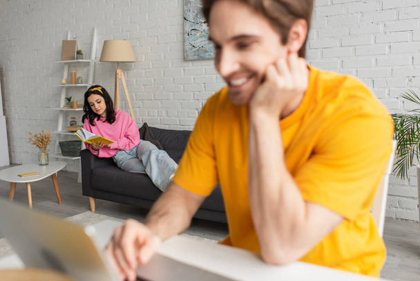 young woman in casual clothes lying with book on couch near blurred smiling boyfriend sitting at table with laptop in living room - Photo, image