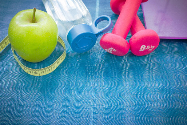 два красных гантели, яблоко, рулетка, бутылка воды и весы на коврике для йоги. концепции фитнеса, спорта и здоровья - Фото, изображение