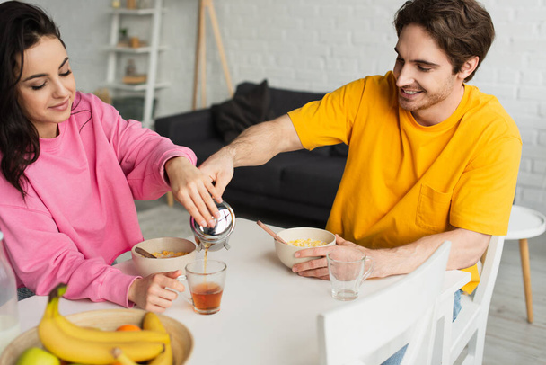 smiling young couple sitting at table with breakfast and pouring tea from french press into cup in living room - Foto, imagen