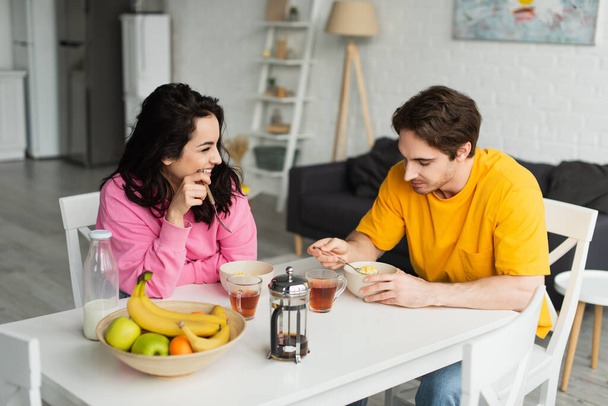 smiling young couple sitting at table and eating breakfast in living room - Foto, immagini