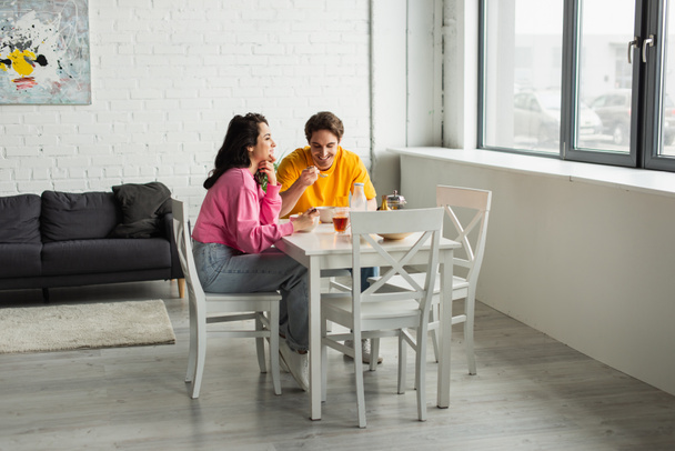 pozytywna młoda para siedzi przy stole przy oknie i je śniadanie w nowoczesnym salonie - Zdjęcie, obraz