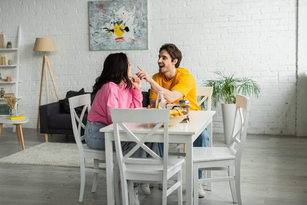 positive young man pointing with finger at nose of girlfriend sitting at table with breakfast in modern living - Photo, Image