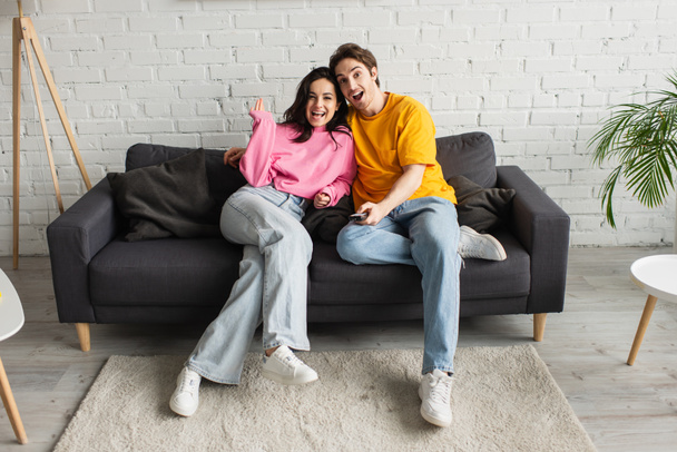 smiling young man pointing with remote controller at camera and hugging girlfriend in living room - Photo, Image